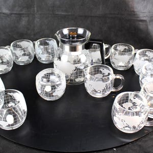 May include: A clear glass coffee set with a world map design. The set includes a coffee maker, creamer, sugar bowl, and several mugs and glasses. The set is arranged on a black surface.