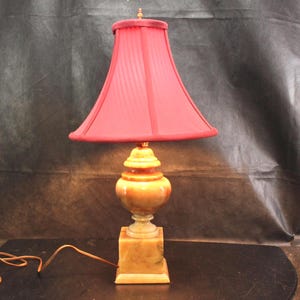 May include: A table lamp featuring a deep red fabric lampshade with a flared shape and a smooth, polished amber-colored stone base. The base consists of a square bottom section, a rounded central body, and a small finial at the top of the shade.