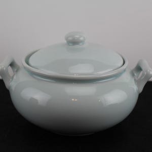 May include: A light blue ceramic serving dish with a lid and two handles. The dish is round and has a smooth, glossy finish.