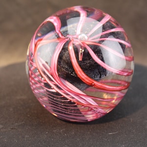May include: A clear glass paperweight with pink and white swirls inside. The swirls are arranged in a spiral pattern and the center of the paperweight is filled with a dark gray substance.