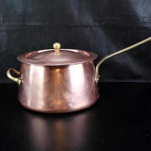 May include: A round copper cooking pot with a matching lid, featuring a long brass handle on one side and a smaller brass loop handle on the opposite side. The pot has a polished metallic finish and a rounded base with a flat rim for the lid.