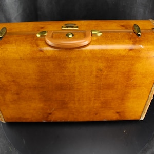 May include: A vintage suitcase with a warm, amber-toned leather exterior. The rectangular case features rounded corners, a brown leather handle, and brass-colored hardware. The suitcase is closed with two latches and a central clasp.