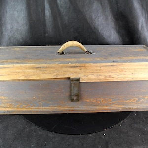 May include: A rectangular wooden tool box with a weathered gray finish and visible wood grain. The box features a hinged lid, a central metal latch on the front, and a curved, light-colored wooden handle attached to the top with metal hardware.