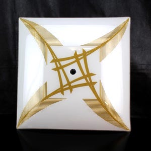May include: A square glass ceiling light shade featuring a white base with a gold-colored graphic pattern. The design consists of four feathered arrow-like motifs pointing toward the corners and a central geometric lattice with a circular hole.