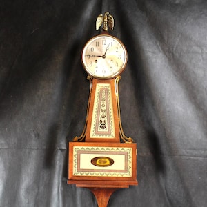 May include: A vintage banjo clock with a round clock face and a decorative eagle finial. The clock has a wooden case with ornate painted details in cream and brown. The clock is approximately 30 inches tall.