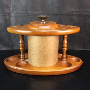 May include: A wooden carousel-style organizer featuring a central cylindrical compartment with a textured gold finish, topped with a small metal knob. The base and top tiers are oval-shaped with carved wooden supports and circular indentations.
