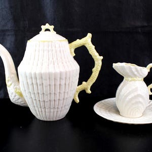 May include: A white porcelain tea set with a textured pattern. The set includes a teapot with a yellow branch-like handle and accents, a creamer, and a saucer. The teapot has a curved spout and a lid with a yellow finial.
