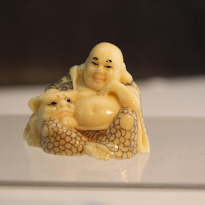 May include: A small ivory figurine of a smiling Buddha sitting with a smaller figure of a dragon in his lap. The Buddha is wearing a robe with a detailed pattern of scales.