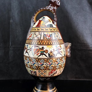 May include: A ceramic vessel with a dark brown base and a pedestal foot, featuring a handle topped with a sculpted feline figure. The body is decorated with colorful geometric patterns, stylized human figures, and llamas, with the text CHINCHEROS CUSCO PERU.