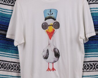 Könnte beinhalten: Weißes T-Shirt mit einem Motiv einer Cartoon-Möwe, die eine Sonnenbrille und einen blauen Matrosenhut mit Anker trägt.