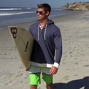 Young man wearing sun glasses and a blue hoodie.  He is carrying a surfboard and walking on the beach in the early morning.