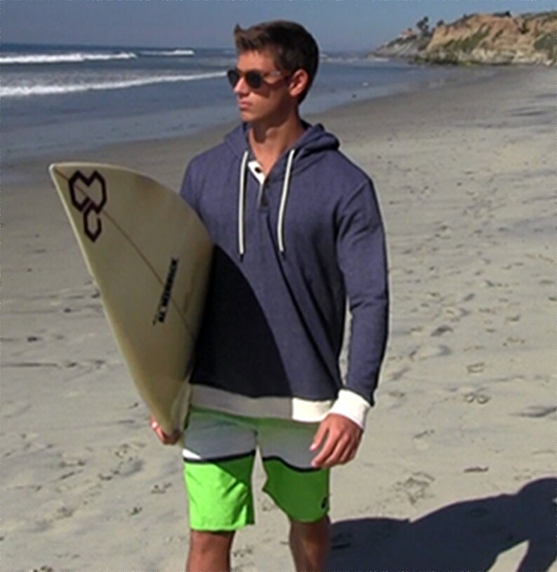 Young man wearing sun glasses and a blue hoodie.  He is carrying a surfboard and walking on the beach in the early morning.