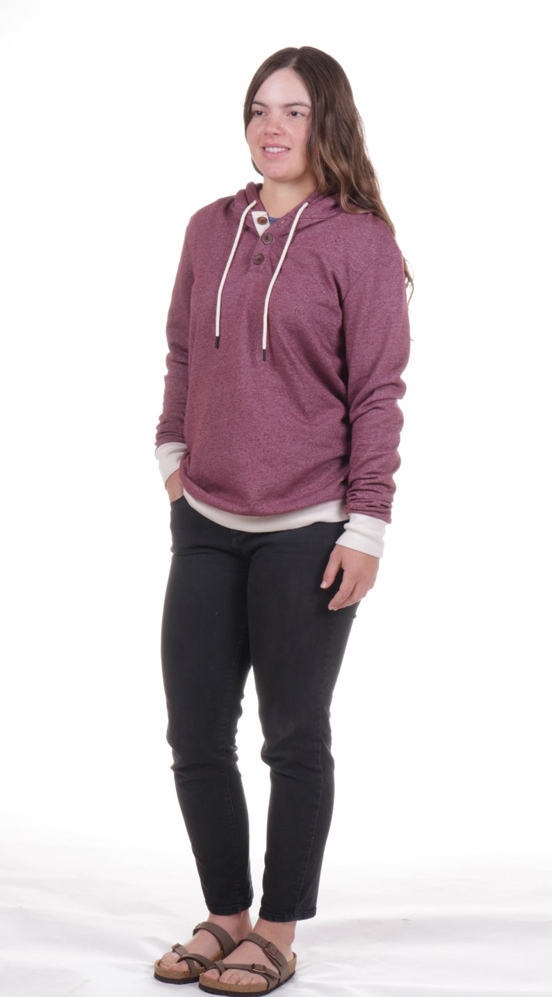 May include: A woman wearing a maroon hoodie with a white trim and a button closure. She is wearing black jeans and brown sandals.