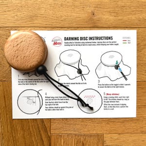 May include: Instructions for using a wooden darning disc to mend fabric. The disc is shown in three stages: 1) with the fabric centred on the disc, 2) with the elastic secured around the disc, and 3) with the fabric secured at the right tension. The instructions include a diagram showing how to make warp stitches.