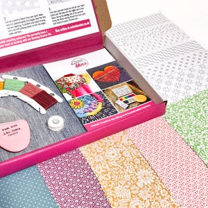 May include: A craft kit box with a pink interior, containing denim fabric, colourful embroidery floss, and a pink needle holder. The box also includes patterned fabric swatches and a booklet with embroidery project images.