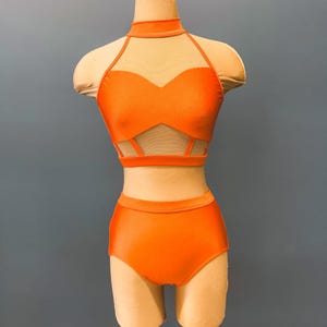 May include: An orange two-piece dance costume displayed on a mannequin. The top features a halter neck, mesh panel, and cut-out design. The high-waisted bottoms complete the set. The costume is made of a shiny, smooth fabric.