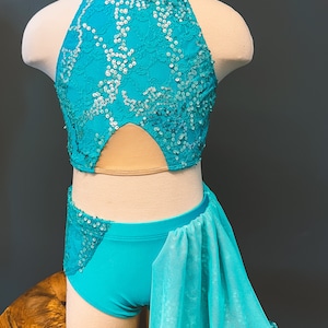 May include: A turquoise dance costume with a halter top and matching shorts. The top is adorned with lace and sequins, and the shorts feature a flowing, sequined skirt overlay. The costume is displayed on a mannequin.