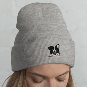May include: A gray beanie with a black and white border collie peeking out from the front. The beanie is made of a soft, knitted fabric.