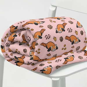 May include: A pink fleece blanket with a repeating pattern of red pandas and brown coffee beans. The blanket is rolled up and placed on a white chair.