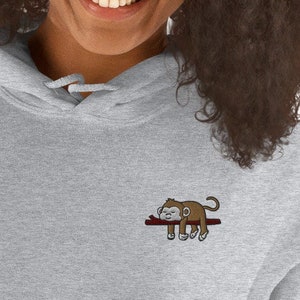 Puede incluir: Una sudadera con capucha gris con una pequeña imagen bordada de un mono durmiendo en una rama.