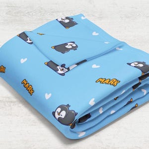 May include: A blue fleece blanket with a repeating pattern of penguins and white hearts. The blanket is folded and the text "MARK" is visible in yellow letters.