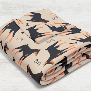 May include: A folded beige fleece blanket with a repeating pattern of black and orange Doberman Pinscher silhouettes and white bone shapes.
