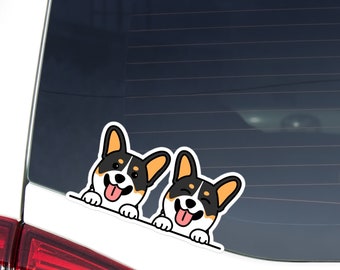 May include: A white sticker featuring two cartoon corgi dogs with black, white, and tan markings. The dogs are peeking over a white edge, with their tongues sticking out and smiling faces. The sticker is likely for a car window.
