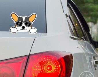 May include: A white car with a cartoon sticker of a corgi wearing sunglasses on the rear window. The corgi is looking at the camera and has its paws on the edge of the window. The car has a red rear light.