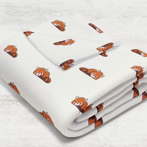 Puede incluir: Manta blanca con un patrón repetido de pandas rojos de dibujos animados en tonos marrones y naranjas. La manta está doblada, mostrando el patrón en la parte superior y los bordes. La manta está hecha de un material suave.