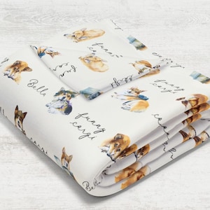 May include: White bedding set featuring watercolor illustrations of corgi dogs in various poses. The words "Bella" and "funny corgi" are written in cursive. The set includes a folded blanket and a pillowcase.