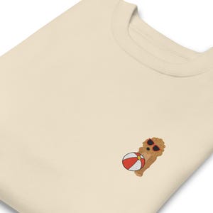 May include: A cream colored sweatshirt with an embroidered image of a dog wearing sunglasses and holding a beach ball.