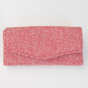 Könnte beinhalten: Eine rot-weiß gewebte Clutch mit Klappenverschluss. Die rechteckige Clutch hat ein strukturiertes Aussehen mit einem Muster aus roten und weißen Fäden. Die Clutch ist geschlossen und liegt flach auf einer weißen Oberfläche.