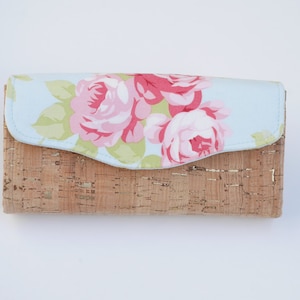Könnte beinhalten: Eine rechteckige Geldbörse aus hellbraunem Kork mit einer hellblauen Klappe, die mit rosa Rosen und grünen Blättern verziert ist. Die Geldbörse hat einen geschwungenen Klappenverschluss. Das florale Muster kontrastiert mit dem Naturkork.