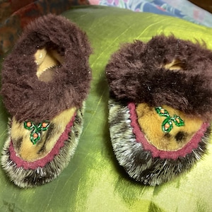 May include: A pair of brown and tan baby moccasins. The moccasins have a dark brown fur trim around the opening and a decorative beaded design on the front. The soles are a mix of brown and gray fur.