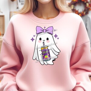 May include: A light pink sweatshirt featuring a cartoon ghost with a purple bow and holding a drink. The drink has the text "Witch's Brew Alan" on it. The ghost is surrounded by purple stars.