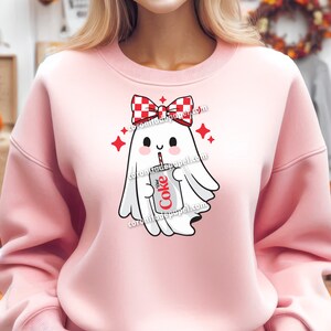 May include: Pink sweatshirt featuring a cute ghost graphic. The ghost wears a red and white checkered bow and holds a Diet Coke can with a straw. Red star accents surround the ghost. The text "Diet Coke" is visible on the can.