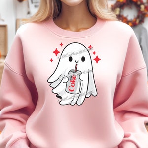 May include: A light pink sweatshirt featuring a cartoon ghost holding a Diet Coke can with a red straw. The ghost is white with a smiling face and surrounded by red star accents. The text "coronitadepapel.com" is printed on the sweatshirt.