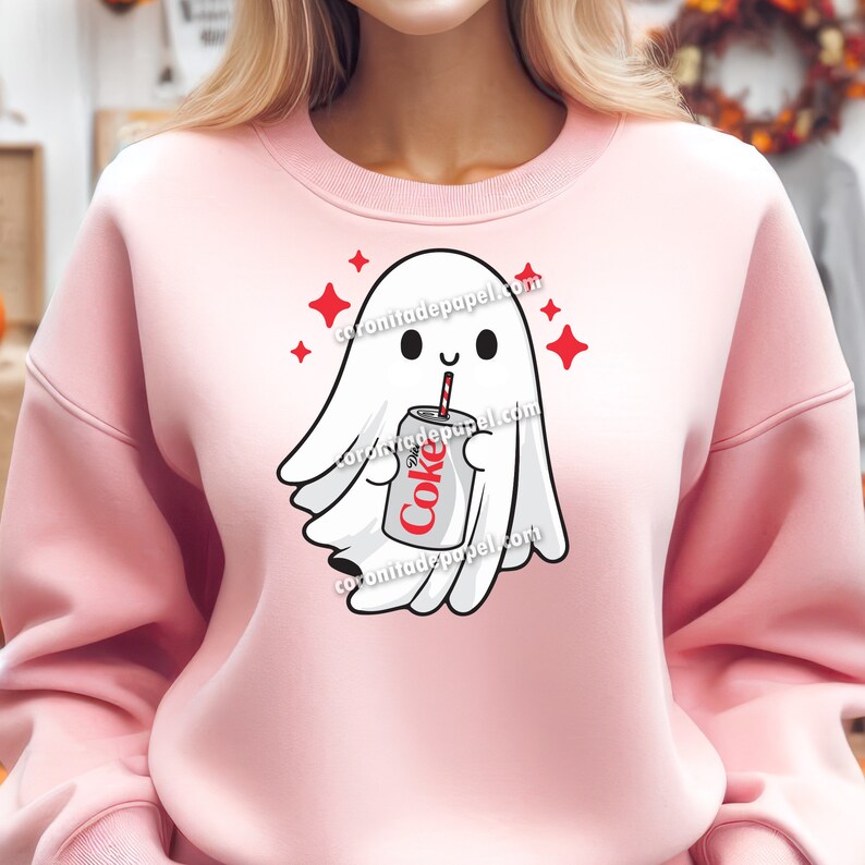 May include: A light pink sweatshirt featuring a cartoon ghost holding a Diet Coke can with a red straw. The ghost is white with a smiling face and surrounded by red star accents. The text "coronitadepapel.com" is printed on the sweatshirt.