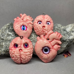 May include: Four pink resin pendants shaped like a brain and a heart, each with a large, colorful eye. The brain pendants have a textured surface, and the heart pendants have detailed anatomical features. Each pendant has a small metal loop.