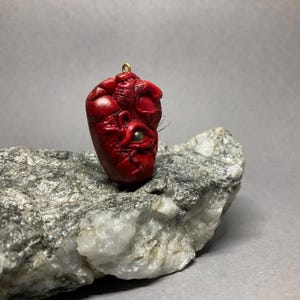 May include: A red heart-shaped pendant with a single eye and gold accents. The pendant is made of polymer clay and is hanging from a gold chain.