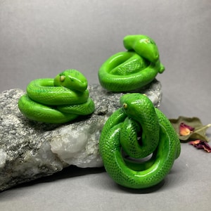 Op de afbeelding: Drie groene keramische slangenbeeldjes, elk op een andere manier opgerold. De slangen zijn lichtgroen met donkergroene markeringen. Ze worden getoond op een grijs en wit rotsoppervlak.