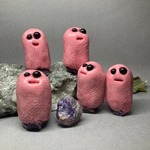 May include: Five pink clay figurines with black eyes and mouths, resembling cartoon characters, are arranged on a rock surface. A purple amethyst crystal is in the foreground.