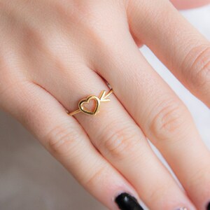 Könnte beinhalten: Ein goldener Ring mit einem Herz- und Pfeil-Design. Der Ring wird an einem Finger mit schwarzem Nagellack getragen.