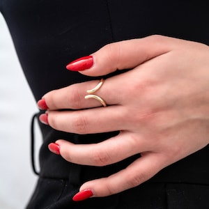 May include: A gold ring with a unique open design. The ring is worn on the middle finger of a hand with red nail polish.