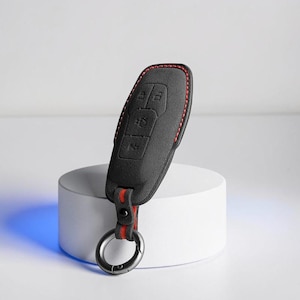 May include: Black key fob cover with red stitching and a red and black strap. The cover has three buttons and a metal key ring. The key fob is displayed on a white cylindrical stand.