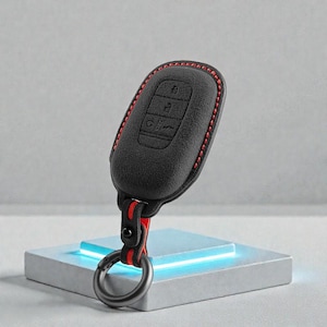 May include: Black key fob cover with red stitching and a red and black strap. The key fob has buttons for locking, unlocking, and other functions. A silver-colored metal ring is attached to the strap.