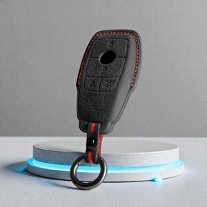 May include: A black key fob cover with red stitching and a red and black strap, attached to a silver key ring. The key fob cover is on a white circular stand with a blue light.