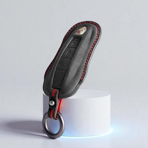 Puede incluir: Una funda negra para llavero con costuras y detalles en rojo. La funda tiene una superficie texturizada y un accesorio para llavero. La funda del llavero se muestra sobre un soporte cilíndrico blanco.