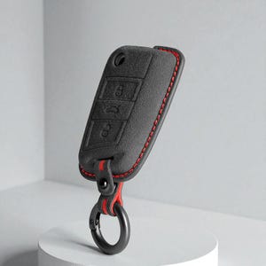 May include: A dark gray textured key fob cover featuring embossed button icons, accented with vibrant red stitching along the edges. The cover includes a matching dark gray strap with red interior lining, secured by a black metal circular carabiner.