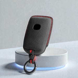 Puede incluir: Una funda negra para llavero con costuras rojas y una correa roja. La funda del llavero tiene un anillo de metal negro para llaves. La funda del llavero está sobre un pedestal blanco.